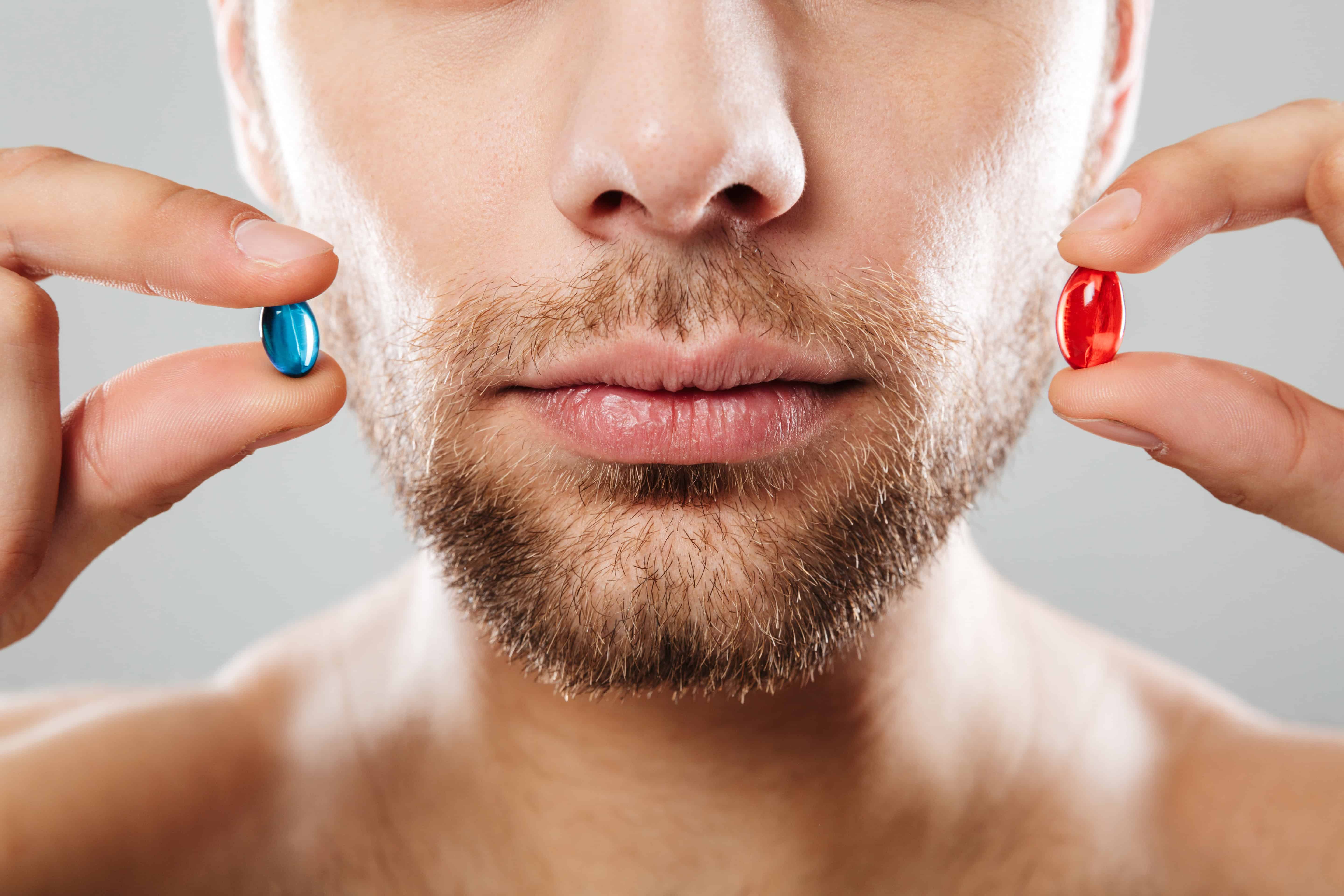 Homme tenant deux pilules, une bleue et une rouge, près du visage.